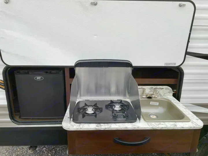 An RV rental's side hatch reveals a practical outdoor kitchen setup: mini-fridge, 2-burner stove, and sink.. Jayco Rocky Mountain 242BHSW 2019