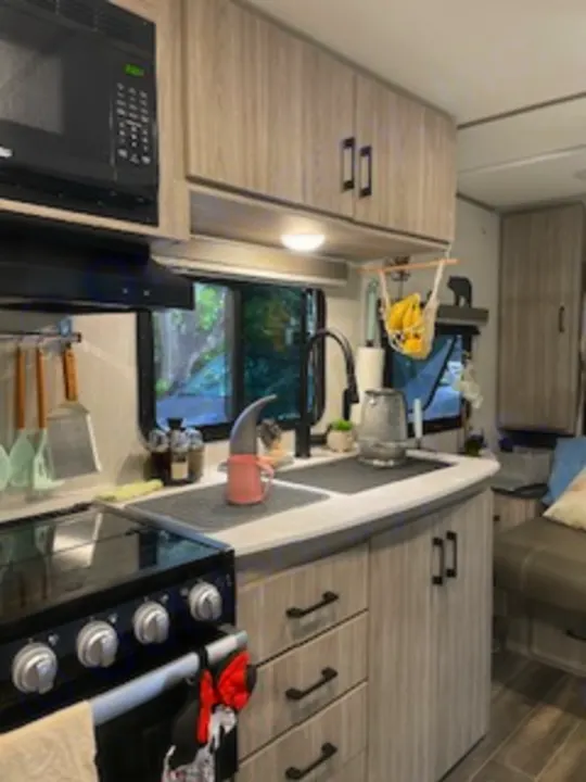 This RV rental kitchen offers compact living with a stove, microwave, sink, and light wood-look cabinets, seen through a window.. Keystone RV Passport SL 2021