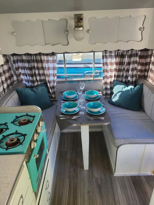 Cozy vintage RV rental interior with a teal kitchen stove, set dinette, comfy bench seating, and charming checkered curtains.. Shasta 1500 1956