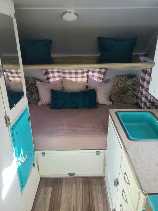 Cozy RV rental interior featuring a full bed with decorative pillows, bright teal sink, and plaid curtains.. Shasta 1500 1956