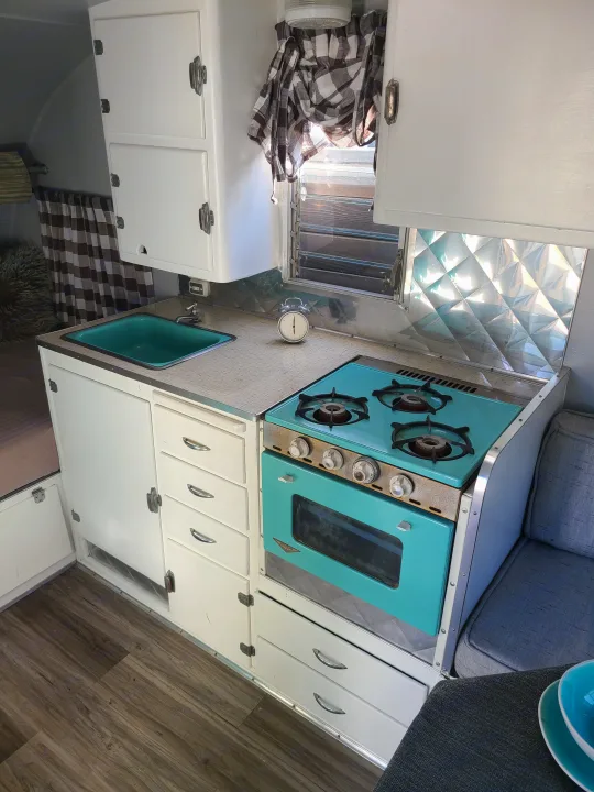 Explore this charming vintage RV rental interior with a vibrant teal kitchen, white cabinets, and wood-look flooring.. Shasta 1500 1956