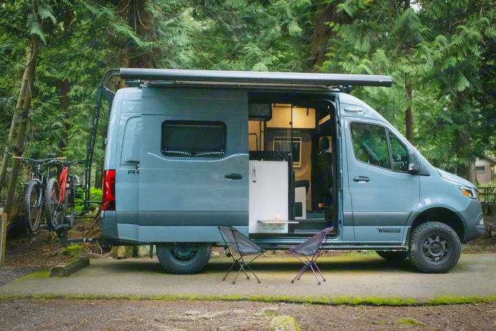 A sleek camper van RV rental with bikes ready for adventure in a tranquil forest campsite, side door open, illuminated inside.. Winnebago Revel 2022