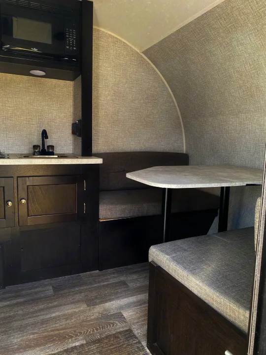 Inside an RV rental: compact kitchen with microwave, sink, and dark cabinets. Dinette has gray upholstered seats.. Braxton Creek Bushwhacker Plus 2022
