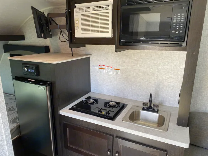 Modern RV rental interior: kitchenette features a two-burner stove, sink, microwave, mini-fridge, AC, and mounted TV.. Braxton Creek Bushwhacker Plus 2022