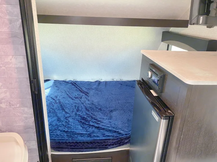 Compact RV rental interior: bed with blue blanket, kitchenette with stereo & mini-fridge, and a partial bathroom view.. Braxton Creek Bushwhacker Plus 2022