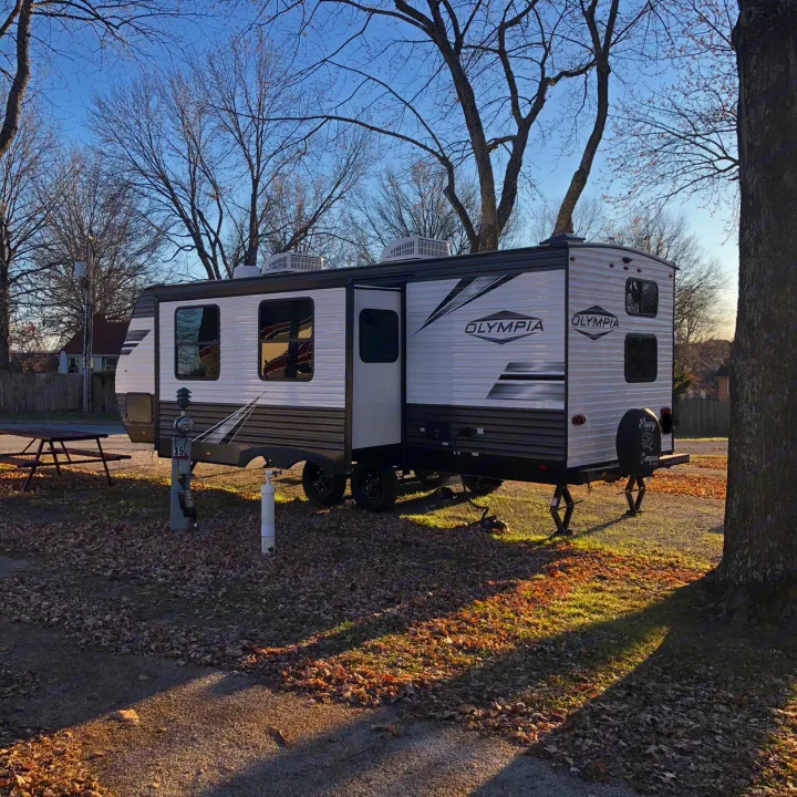 Happy camper! An Olympia travel trailer RV rental is set up at a sunny, leaf-covered campsite, perfect for an autumn getaway.. Highland Ridge Olympia 26BHS 2022