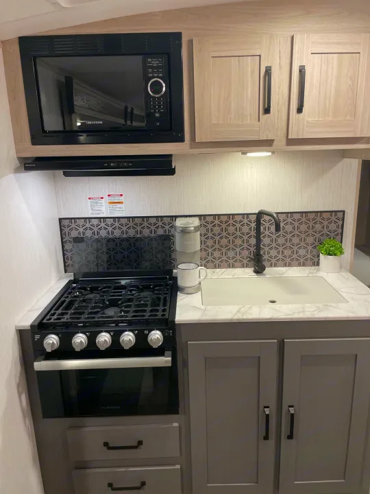 This modern, compact RV kitchen boasts a microwave, gas stove, sink, and stylish backsplash—perfect for an RV rental.. Heartland Sundance 294BH 2022