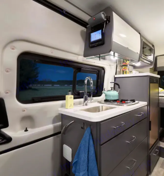 Inside this RV rental: a modern kitchen with sink, stovetop, and storage. The window reveals a dusk field, ideal for camper van travel.. Thor Tranquility 2022