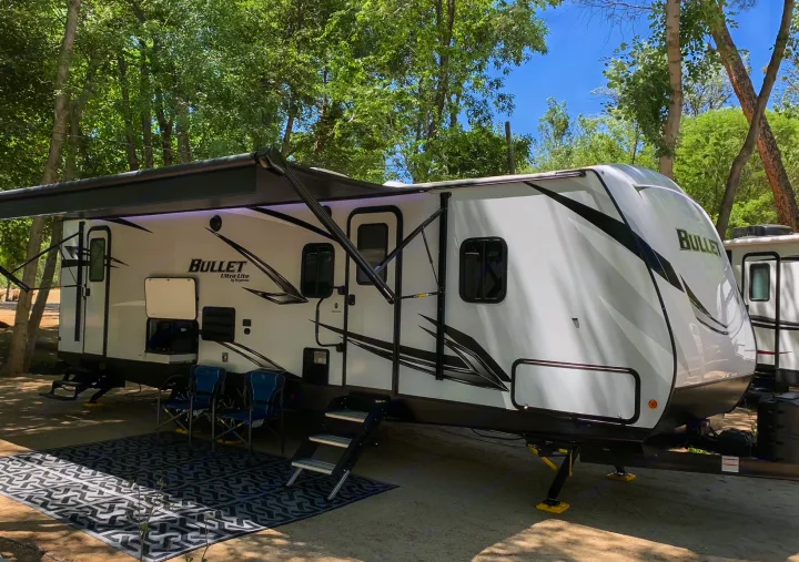 This white Bullet Ultra Lite RV rental is fully set up with an awning, chairs, and outdoor rug at a shady campsite.. Keystone Bullet 2021