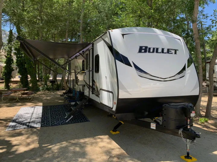 A white Bullet RV rental is seen at a shaded campsite with its awning extended and an outdoor mat laid out.. Keystone Bullet 2021