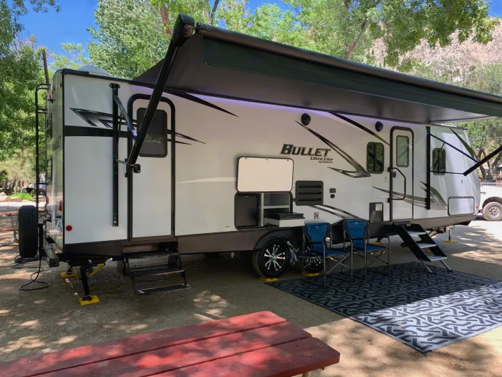 This RV rental travel trailer is perfectly set up at a shady campsite with an awning, chairs, and mat for outdoor enjoyment.. Keystone Bullet 2021