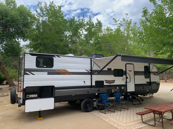 This Wildwood travel trailer, perfect for an **RV rental**, is set up at a shady **campground**, featuring an outdoor living area.. Forest River Wildwood 2021