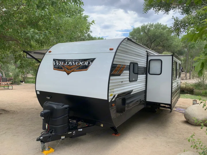 This white and black Forest River Wildwood travel trailer, an ideal RV rental, is set up at a sandy, tree-lined campsite.. Forest River Wildwood 2021