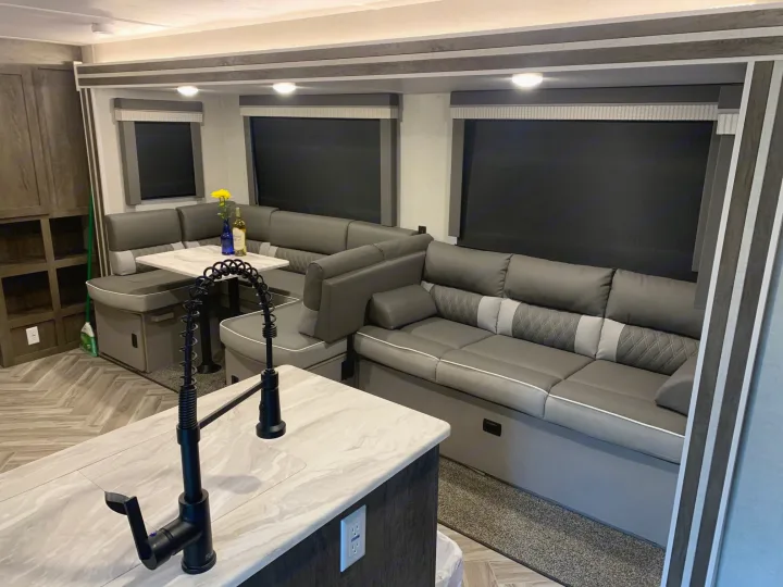 Modern RV rental interior: gray L-shaped sofa, dining table, kitchen island, and window blinds. Cozy and functional space.. Forest River Wildwood 2021