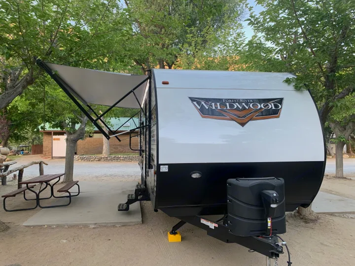 White and black Forest River Wildwood RV rental parked at a shady campsite with an extended awning and picnic table.. Forest River Wildwood 2021
