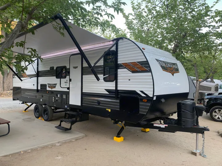 Modern travel trailer RV rental with awning extended, stabilized by yellow blocks, at a shaded campsite.. Forest River Wildwood 2021
