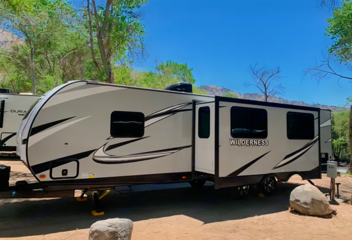 This RV rental, a Wilderness travel trailer, is set up at a sunny outdoor campsite surrounded by trees and mountains.. Heartland Wilderness 2020