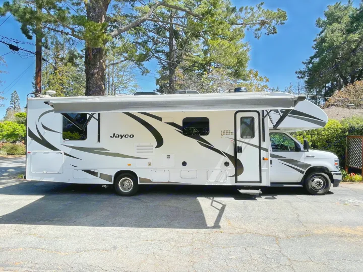 White Jayco Class C RV rental with extended awning. Parked on asphalt under sunny skies, trees in background.. Jayco Redhawk 2021