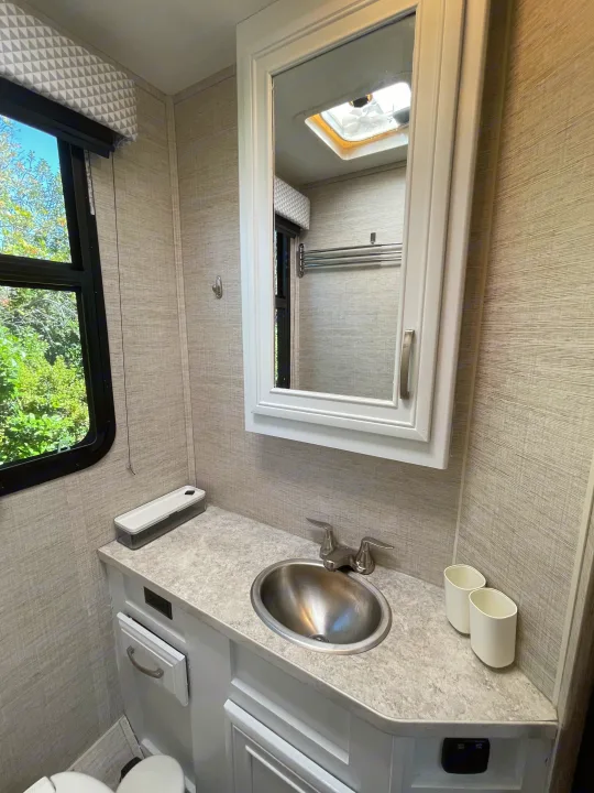 Clean RV rental bathroom featuring a stainless steel sink, mirror, modern gray walls, and bright window views.. Jayco Redhawk 2021