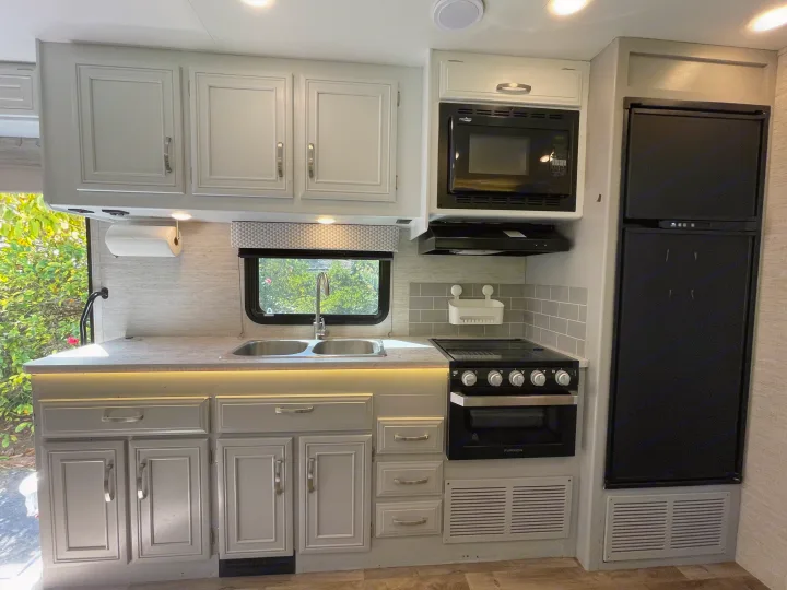 Spacious RV rental kitchen with light gray cabinets, sink, microwave, oven, and fridge. Bright under-cabinet lighting.. Jayco Redhawk 2021