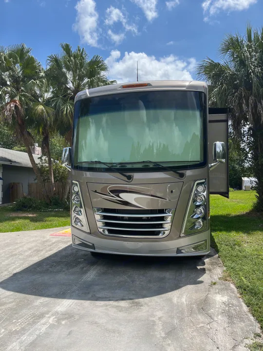 Front view of a sleek tan RV on a sunny driveway, surrounded by palm trees.. Thor Challenger 2021