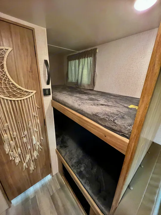Cozy bunk beds inside an RV rental, featuring soft grey bedding, a window with curtains, and a door with moon decor.. Winnebago Micro Minnie 2021