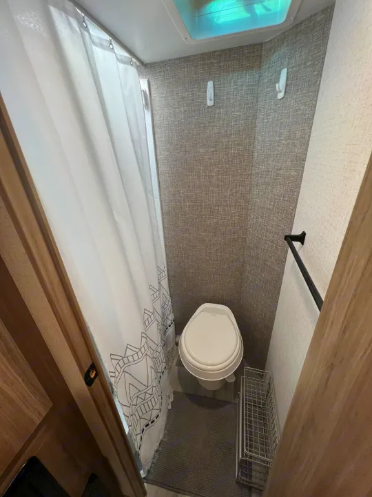 High-angle view of a compact RV rental bathroom featuring a toilet, patterned shower curtain, ceiling vent, and storage.. Winnebago Micro Minnie 2021