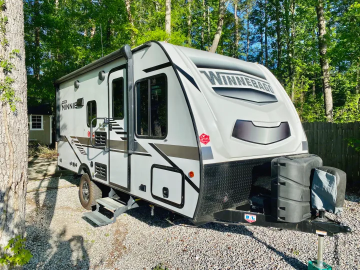 A white Winnebago Micro Minnie travel trailer is ready for an rv rental adventure, parked on gravel beneath green trees.. Winnebago Micro Minnie 2021