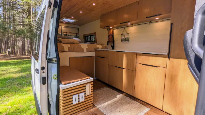 Spacious camper van interior with an open door revealing a forest view. Bed, kitchen, and seating are ready for RV rental.. Mercedes-Benz Sprinter 2020