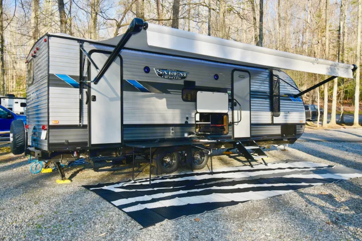 An RV rental, a grey travel trailer with an extended awning and striped mat, set up at a gravel campsite among trees.. Forest River Other 2022