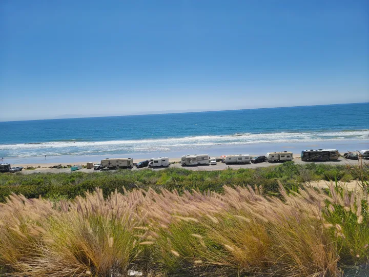 Tall grass frames RVs (motorhomes, trailers) parked by a sandy beach and ocean. Great for an rv rental.. Thor Axis 2021