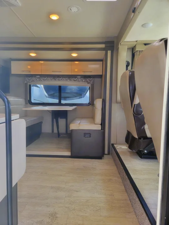 Clean RV rental interior featuring overhead cab-over seating, light-toned wood-look flooring, modern dinette, and bright cabinets.. Thor Axis 2021