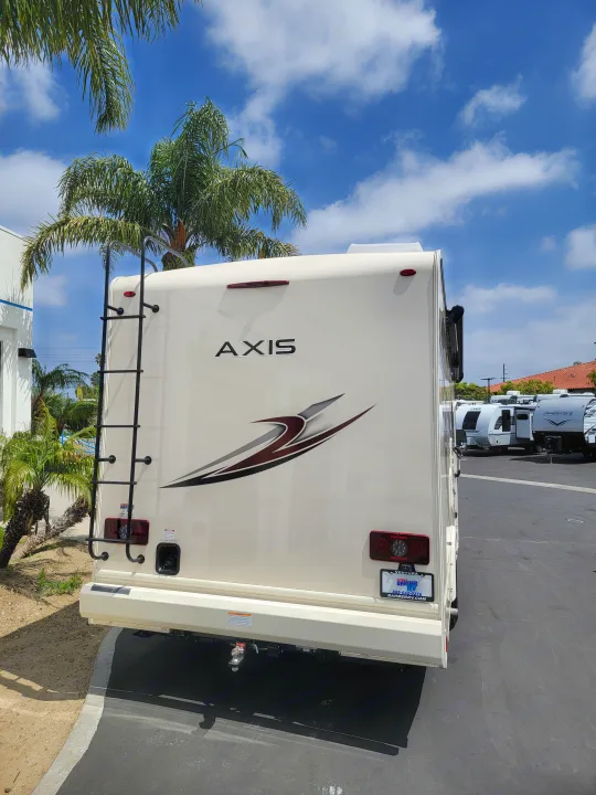Rear of a white Axis RV with a ladder & tail lights, parked outdoors under a blue sky. Ideal for your next RV rental.. Thor Axis 2021