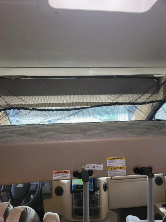 Inside an RV rental: electric overhead bunk with safety net and caution labels visible, showing part of the cockpit.. Thor Axis 2021