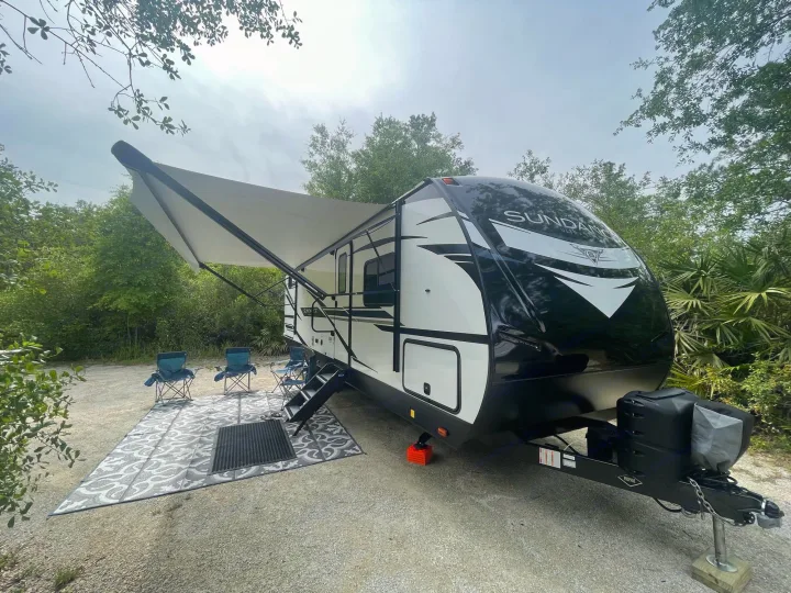 Spacious RV rental set up at a peaceful campsite with an extended awning, outdoor mat, and chairs. Perfect for a nature getaway.. Heartland Sundance 294BH 2022