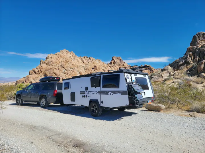 Off-road RV rental: Dark SUV tows a white Mantis travel trailer through rocky desert landscape under a clear blue sky.. TAXA Habitat Mantis 2021