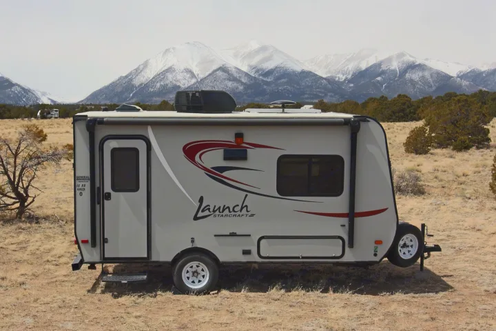 A gray Launch Starcraft travel trailer, ideal for rv rental, rests in a dry landscape with majestic snow-capped mountains.. Starcraft Launch 2016