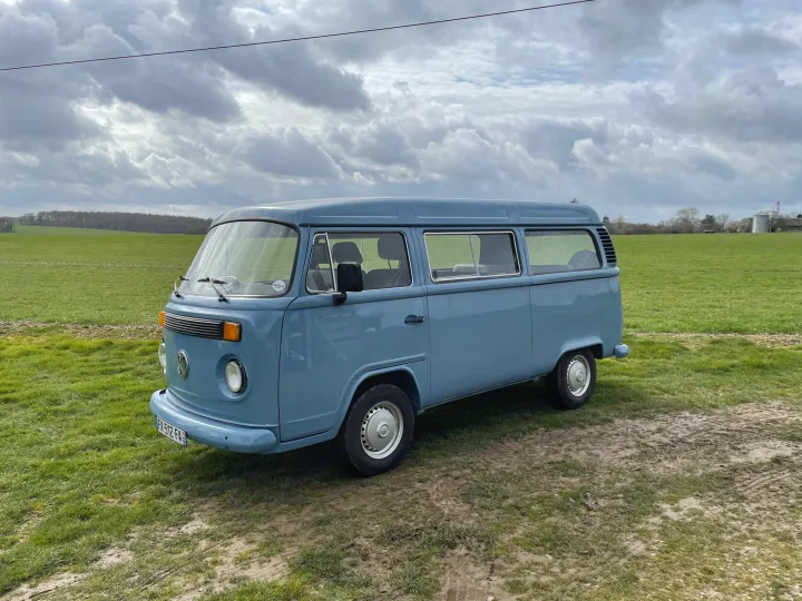 Light blue classic VW camper van on a muddy track in a green field under cloudy skies, perfect for an RV rental adventure.. Volkswagen T2 1980