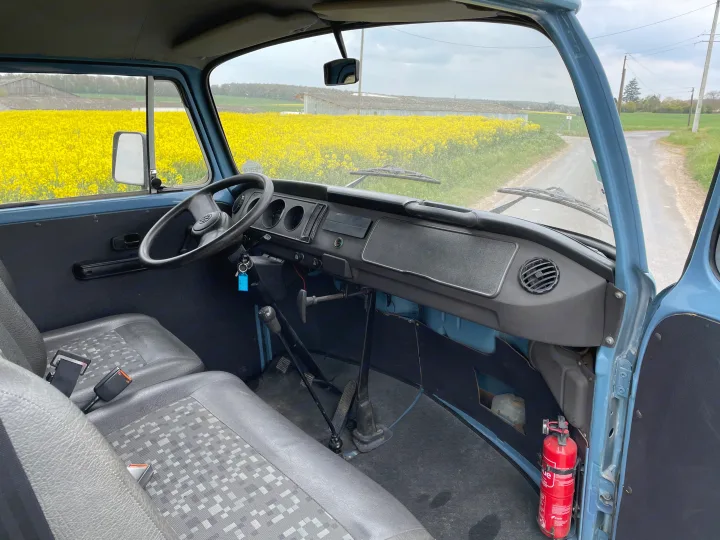 Vintage RV rental dashboard & driver's seat. Outside, bright yellow fields and a dirt road unfold.. Volkswagen T2 1980