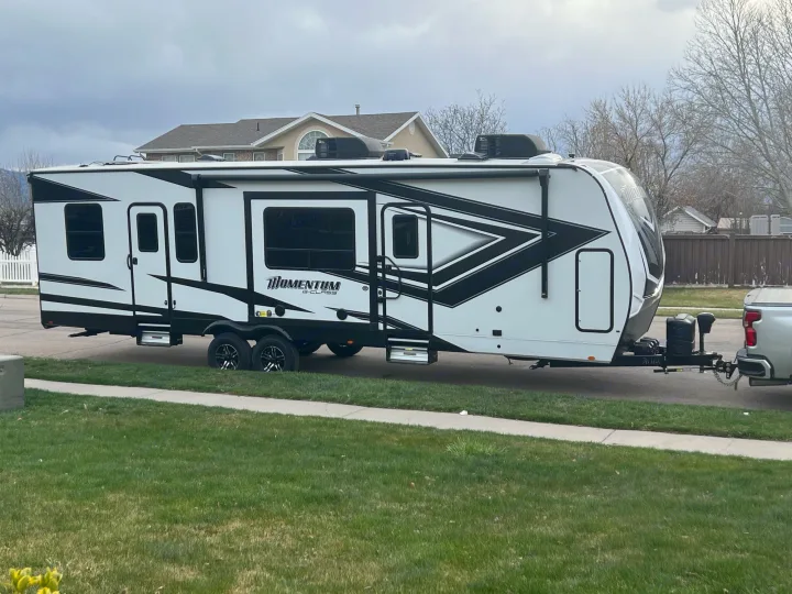 Large white RV with black accents, hitched to a truck & parked on a street, ready for your rv rental adventure.. Grand Design Momentum 2022