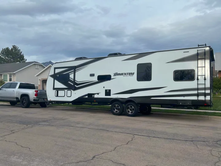 Large white Grand Design Momentum RV toy hauler towed by a silver pickup truck on a street, ready for your rv rental adventure.. Grand Design Momentum 2022