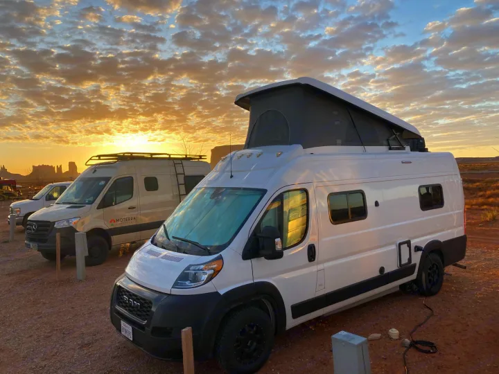 White pop-top campervan & tan RV rental parked in desert at stunning sunset with golden clouds. Epic van life adventure.. Winnebago Solis 59PX 2022