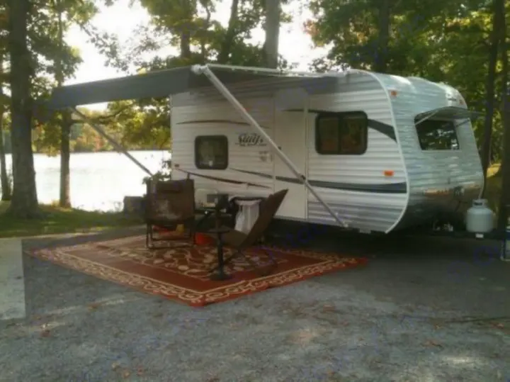 RV rental at a scenic lakeside campsite. An extended awning covers an outdoor rug, chairs, and a fire pit.. Jayco Bambi 2014