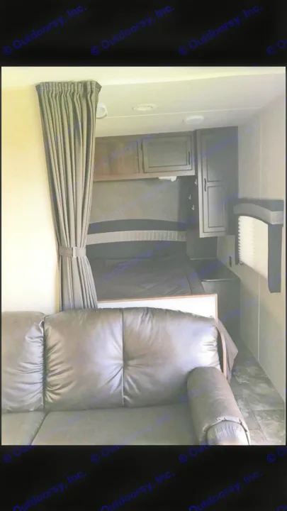 RV rental interior: A dark leather sofa and compact bedroom area with tied-back privacy curtain, bed, and cabinets.. Jayco Coyote 2014