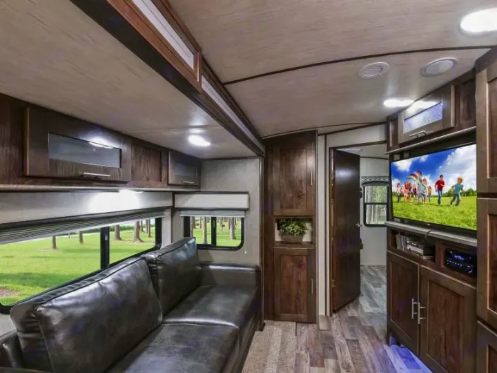 Cozy RV rental living space featuring a dark leather sofa, TV, and ample wood cabinetry. Windows show greenery.. Radiance Big Bear 2020