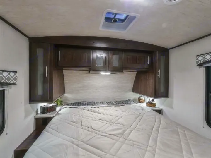 This RV rental bedroom features a queen bed centered under dark wood overhead cabinets. A skylight is visible on the ceiling.. Radiance Big Bear 2020
