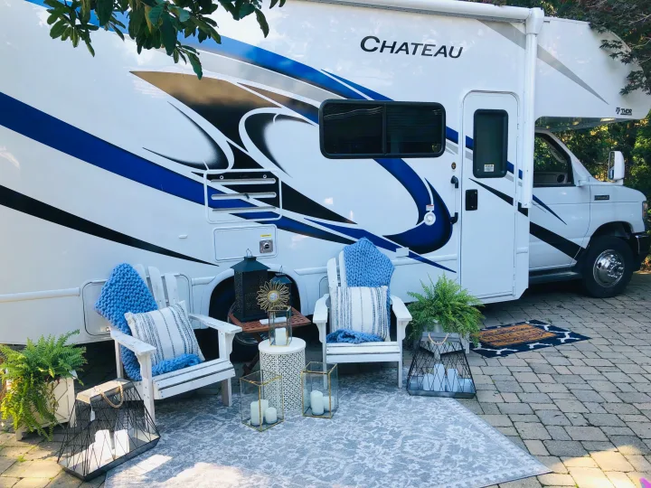 This RV rental offers a stylish outdoor patio with white chairs, a cozy rug, and decorative lanterns next to the motorhome.. Thor Chateau 2022