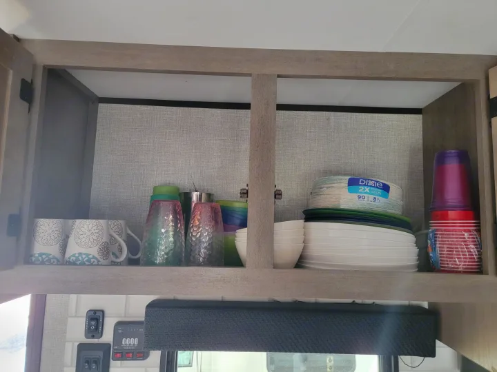 Overhead cabinet in an RV rental, stocked with various reusable and disposable dishes like plates, mugs, and plastic cups.. Jayco Rocky Mountain Edition 2022