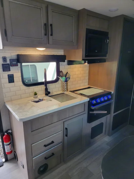 Modern RV rental kitchen: microwave, blue-lit stove, sink, and gray cabinetry. Perfect for comfortable travel and cooking.. Jayco Rocky Mountain Edition 2022