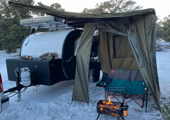 Cozy winter camping with an RV rental teardrop trailer, extended awning, and warm fire pit in the snow.. Bean Mean Bean 2022